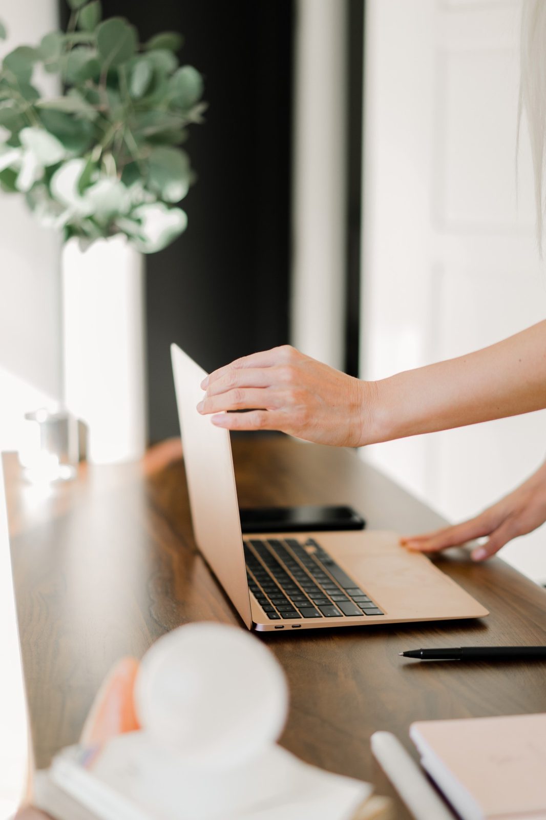 A hand opening a laptop on a walnut desk beside a vase of eucalyptus